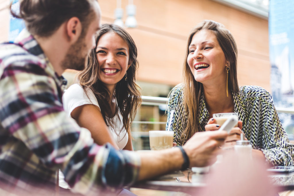 Bilde av en mann og to kvinner rundt et bord. De to kvinnene smiler bredt mens de ser bort på mannen. De ser ut til å ha et kaffemøte.