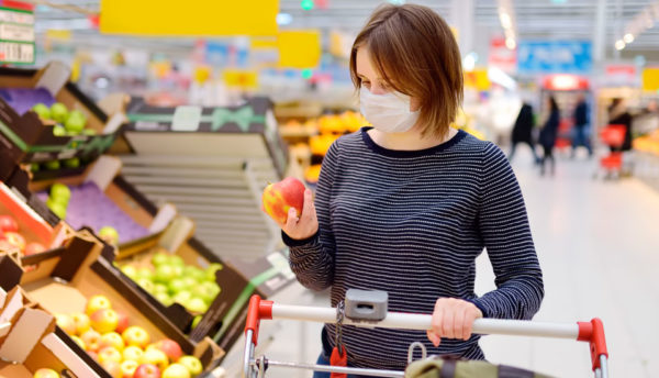 Bilde av en kvinne iført munnbind i fruktavdelingen i en matvarebutikk. Hun hviler den ene hånden på handlevognen og i den andre hånden holder hun et eple.