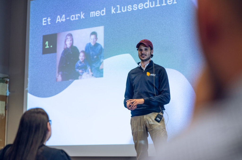 Smilende mann med caps står på scenen og holder et foredrag. I forgrunnen ser vi bakhodet til en kvinne.